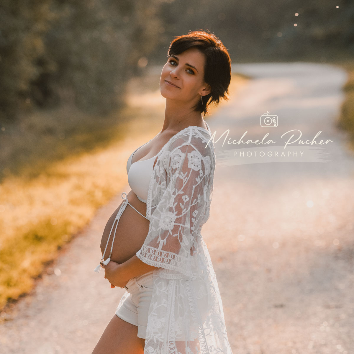 Schwangere Frau im Abendlicht, die seitlich auf einem sonnigen Waldweg steht und liebevoll ihren Babybauch hält.